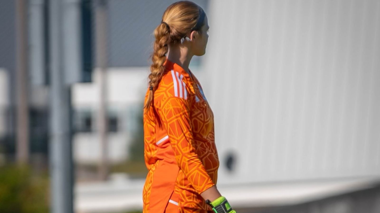 Girls soccer player goalkeeper looks off into the distance during a soccer match, wearing an orange jersey and yellow goalkeeping gloves.