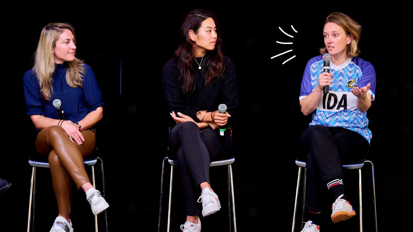 IDA co founder Laura Youngson sits on a stool on stage speaking into a microphone alongside 2 other female founders.