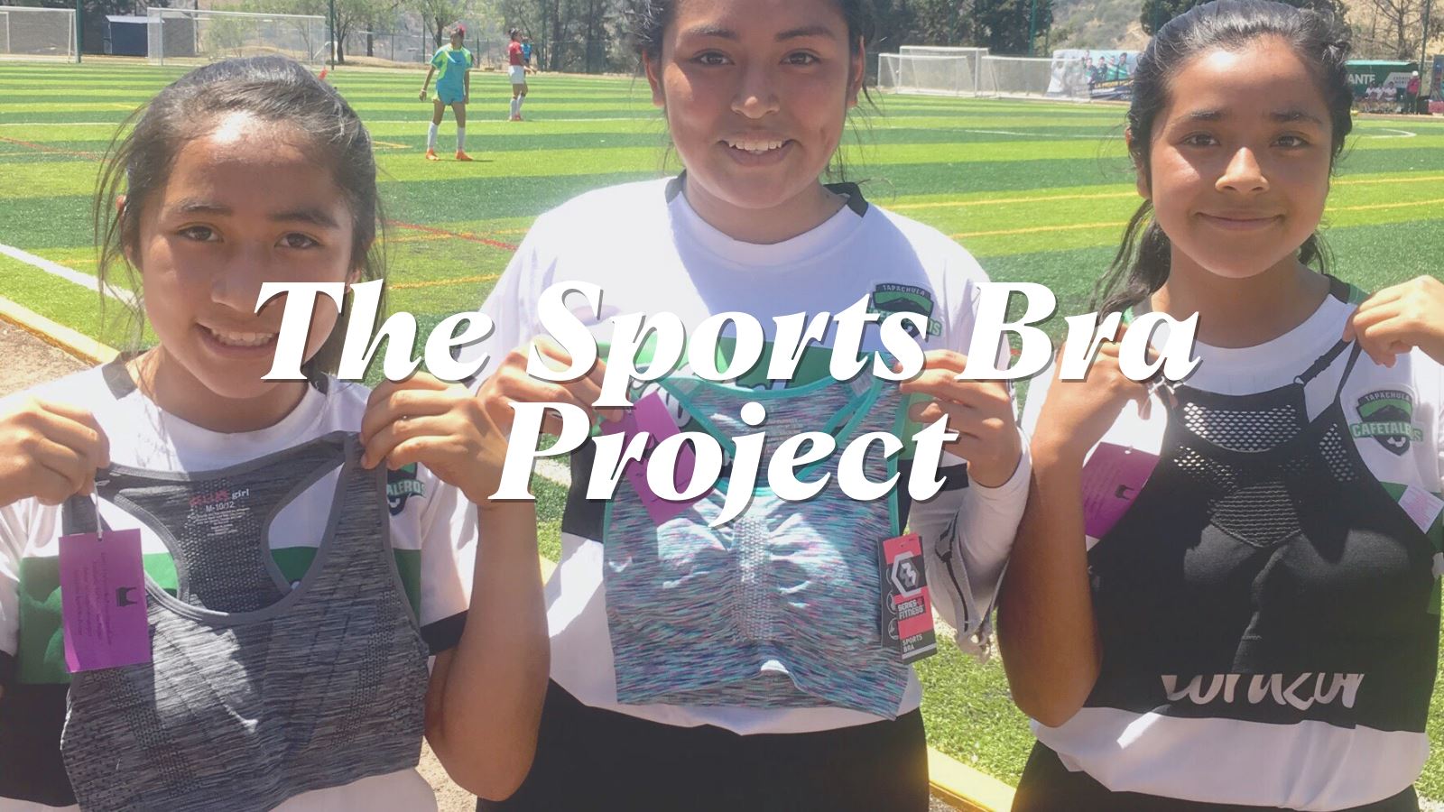 3 female soccer players hold up sports bras smiling at a soccer field. Text says "The Sports Bra Project".