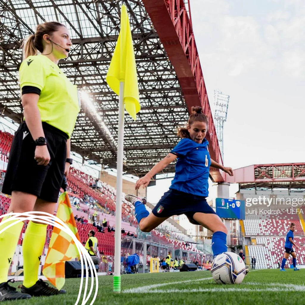 Professional referee Vikki Allan wears the IDA Classica black leather women's soccer cleats while standing at the corner flag and a women's soccer player for Italy takes a corner kick.