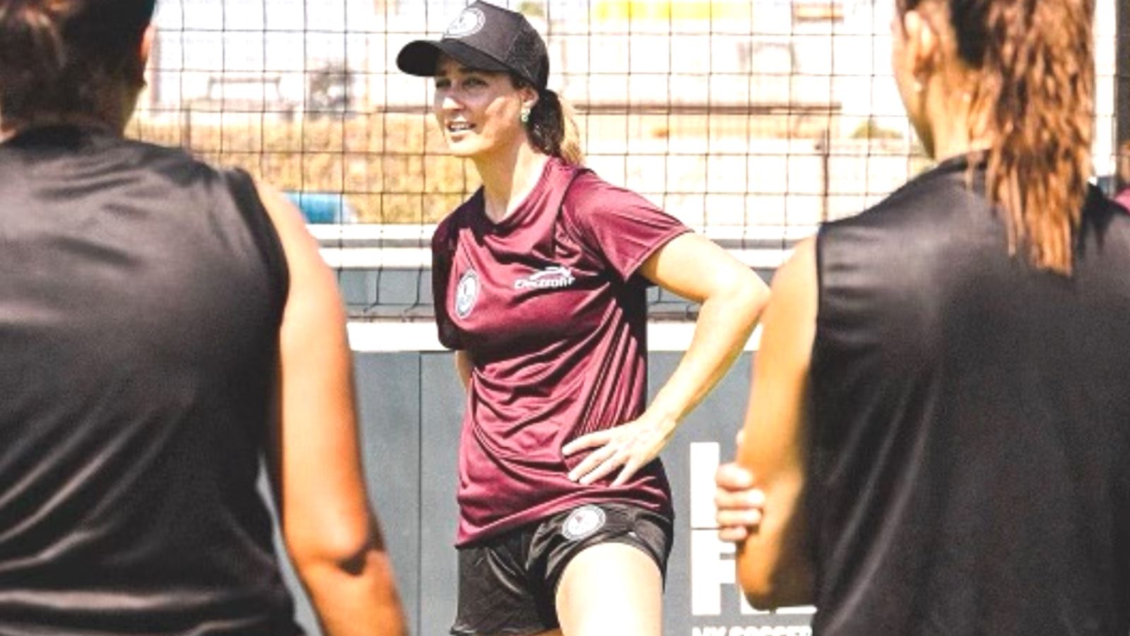 Dr. Nicole Surdyka wearing a tshirt, shorts, and black cap, stands on a soccer field coaching female soccer players.
