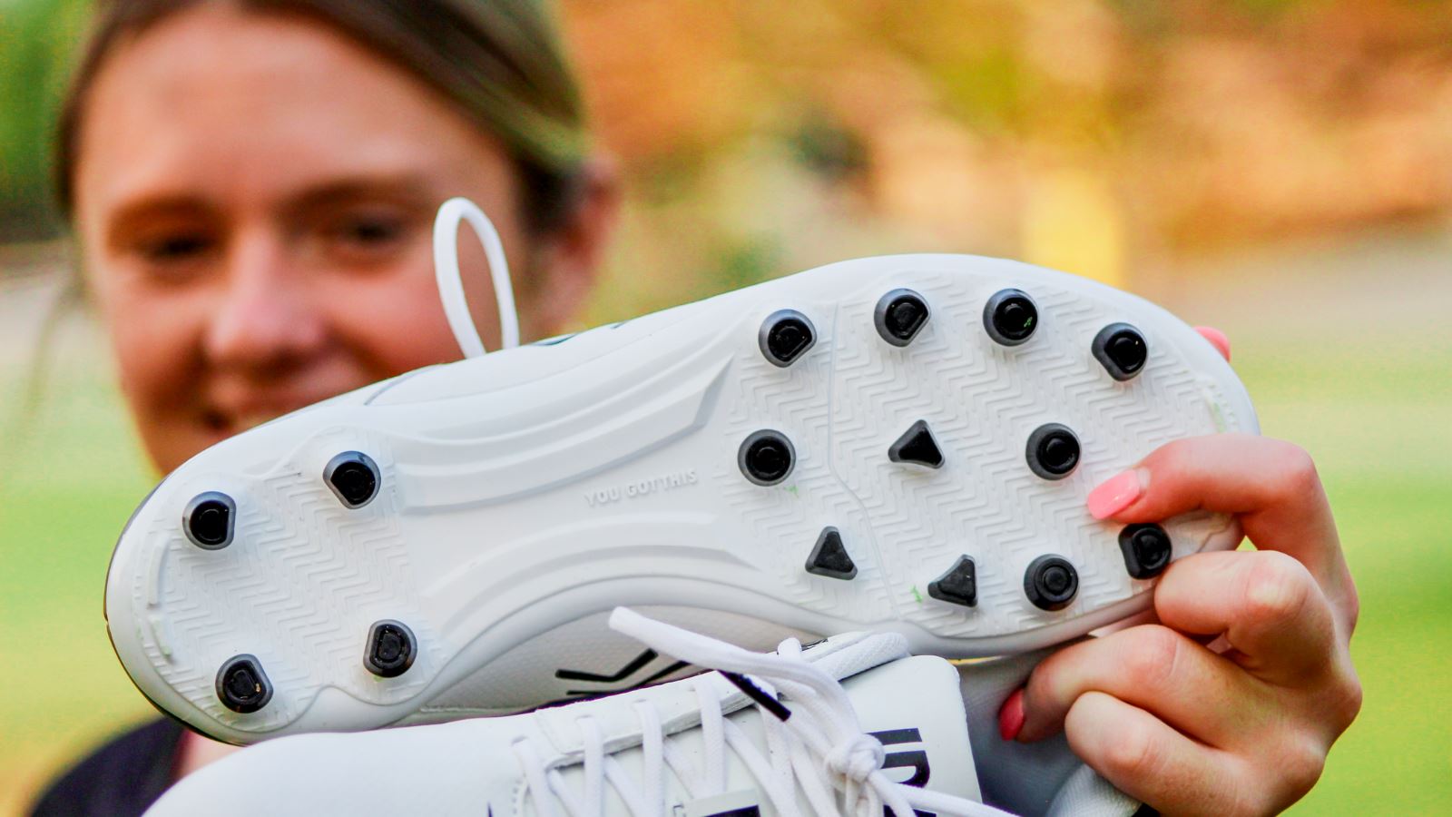 Teenage girl smiles and holds a pair of white IDA Centra women's soccer cleats up in front of the camera with black studs facing up.