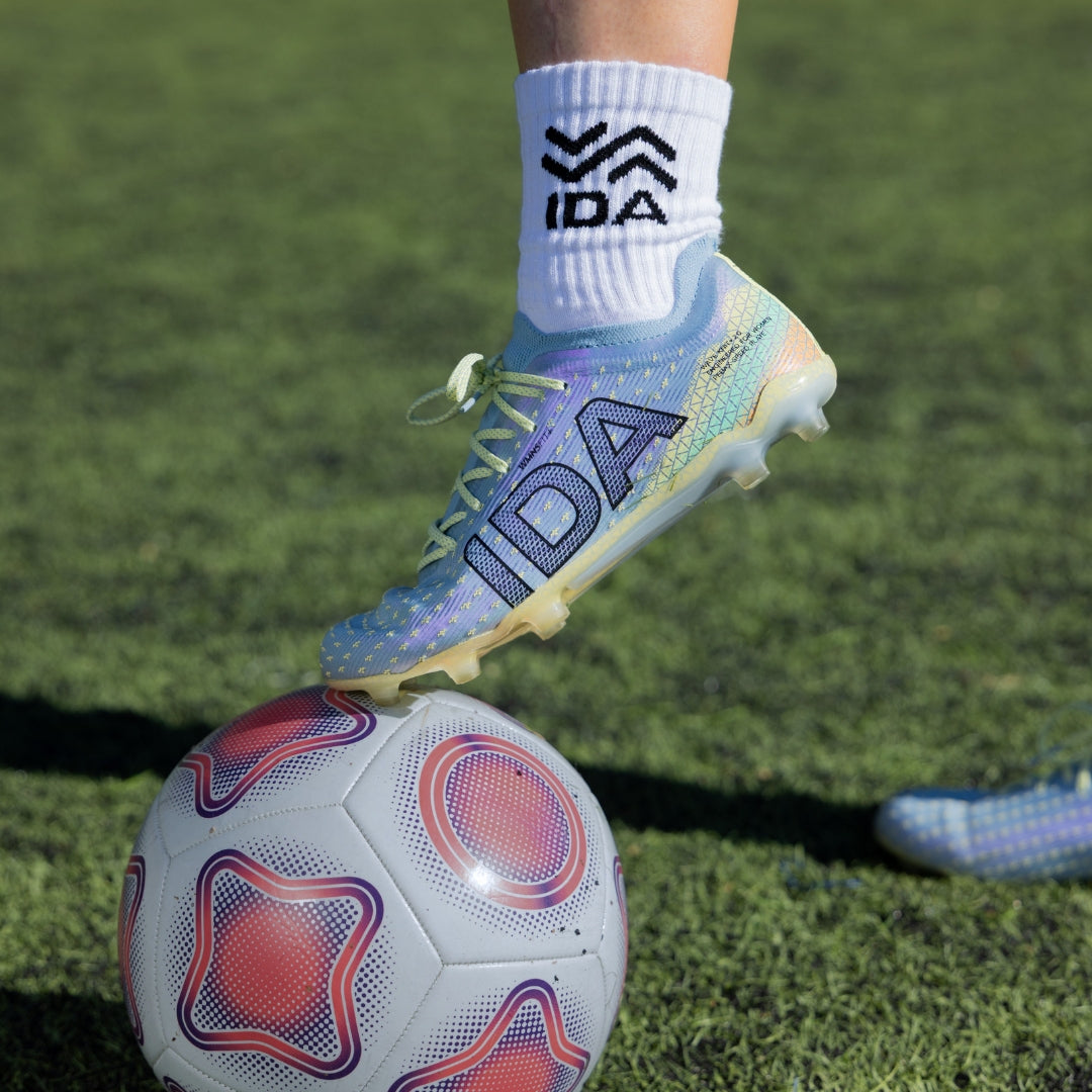 Soccer player's foot wearing a blue cleat with 'IDA' branding on a soccer ball on grass.