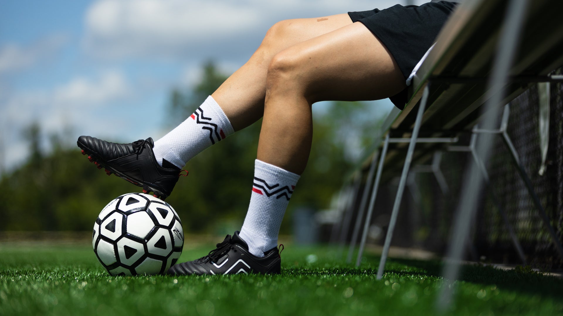 Women's soccer player sits on a bench at a field wearing the IDA Classica black leather women's soccer cleats and resting her feet on a soccer ball.